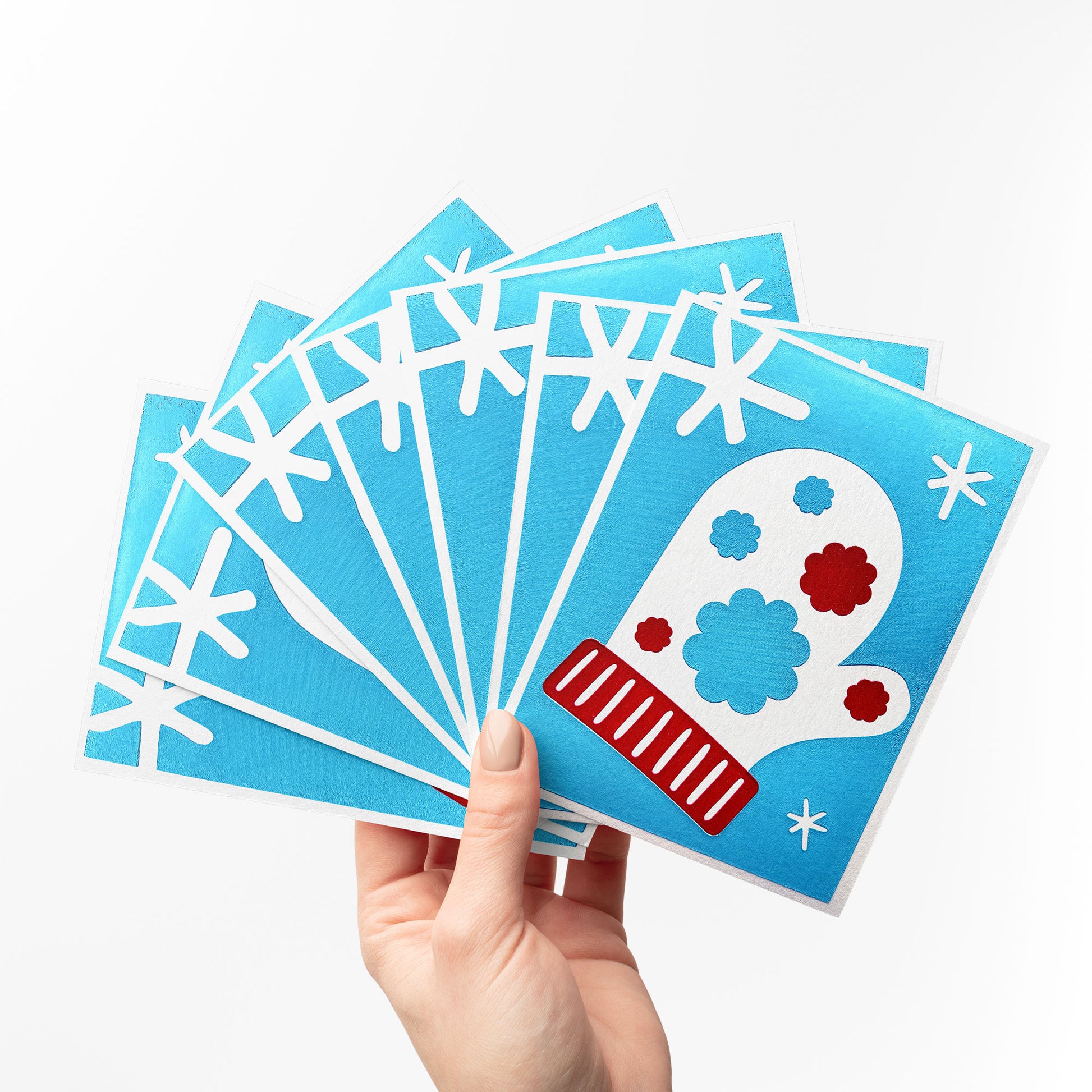 A hand holding a fan display of multiple Snow Blossom Mitten Cards, each featuring a white mitten with red and blue floral accents surrounded by white snowflakes on a vibrant blue background.