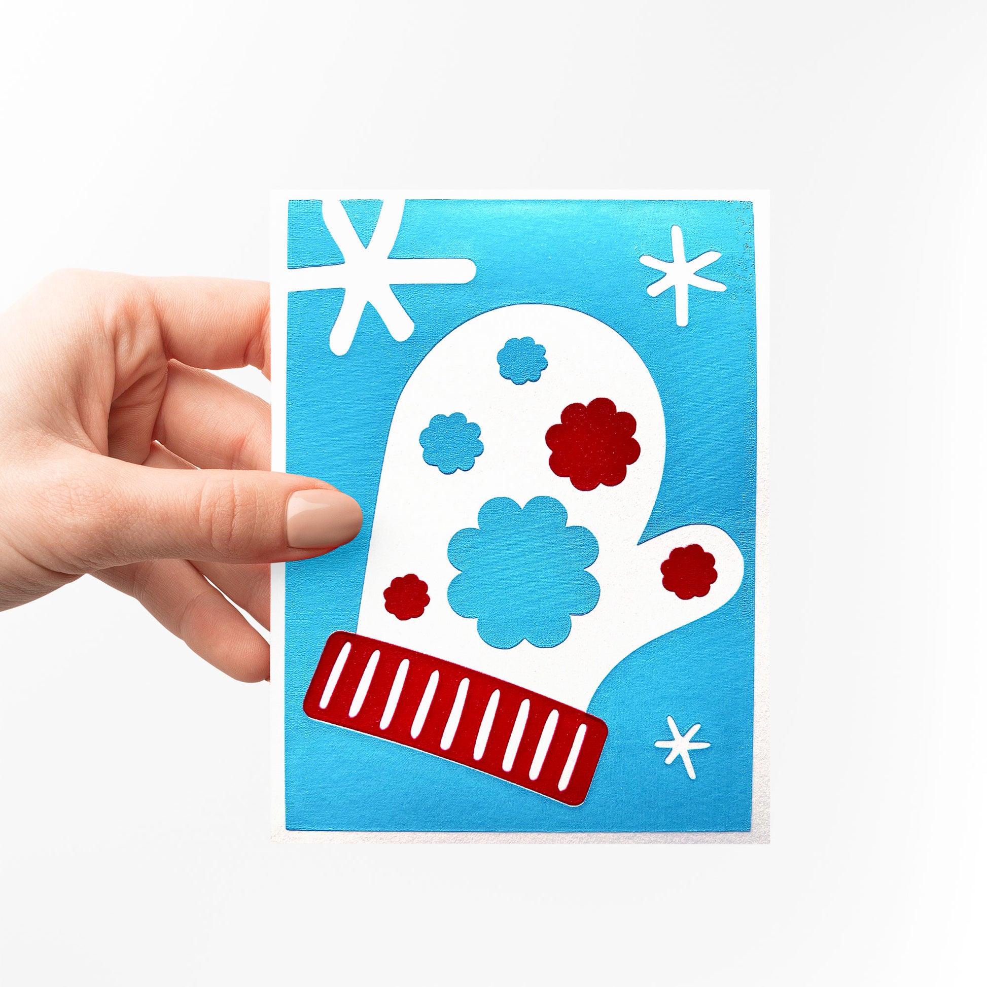 A hand holding the Snow Blossom Mitten Card, a handcrafted winter-themed greeting card featuring a white mitten with red and blue floral accents, surrounded by snowflake designs on a vibrant blue background.