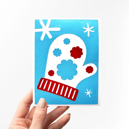 A hand holding the Snow Blossom Mitten Card upright, showcasing its winter-inspired design with a white mitten adorned with red and blue floral accents and surrounded by snowflakes on a bright blue background.