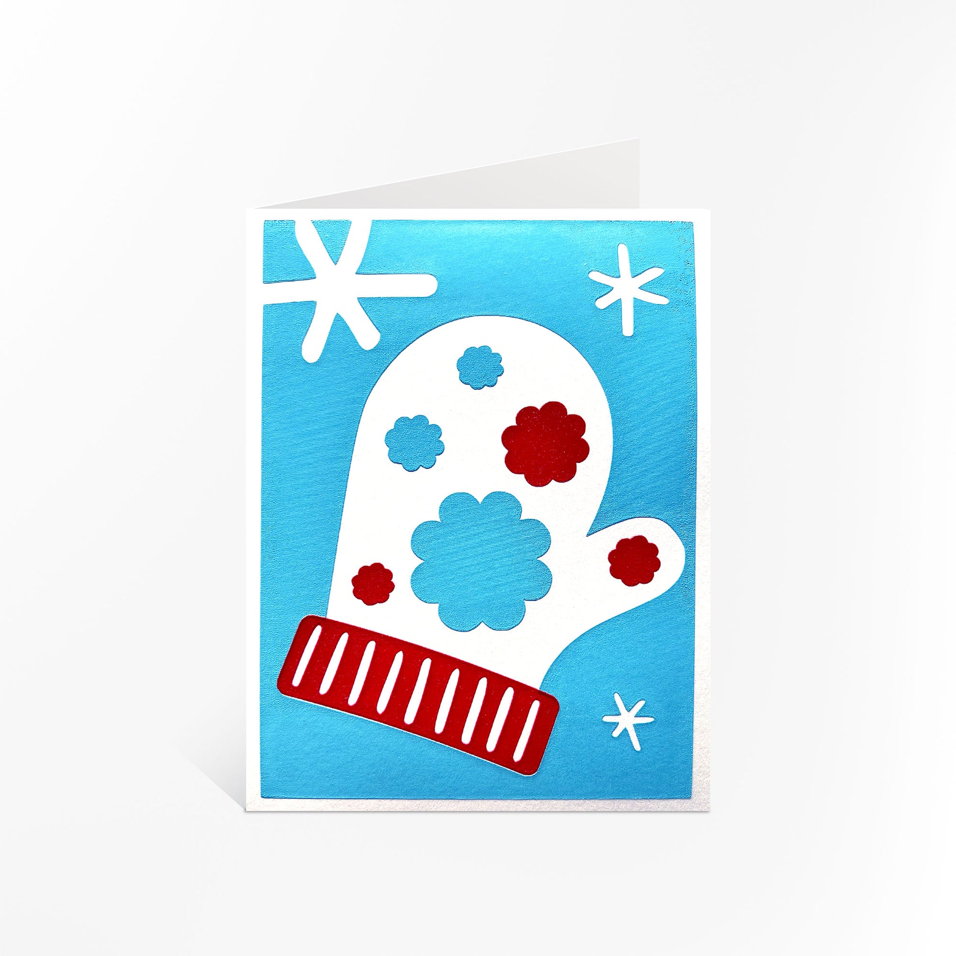 The Snow Blossom Mitten Card displayed standing upright, showcasing its winter-themed design with a white mitten adorned with red and blue floral accents and surrounded by white snowflakes on a vibrant blue background.